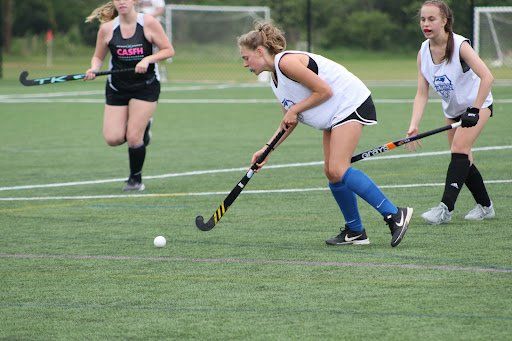 Field hockey players chasing a ball on a green turf field, with one dribbling in the foreground