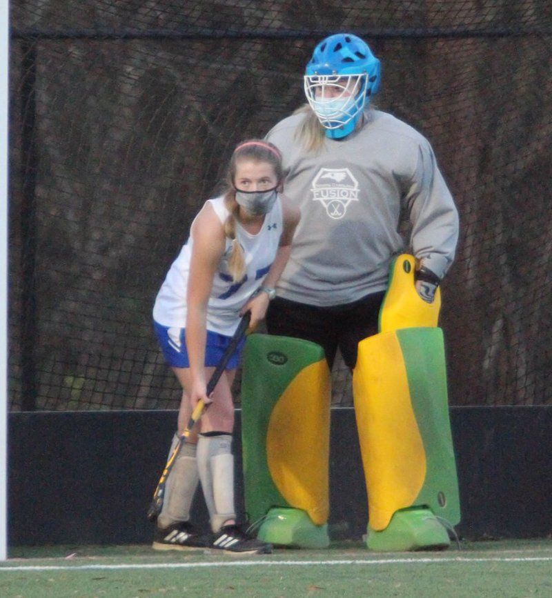 Field hockey players in protective gear stand ready on the turf near the goal