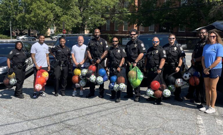 Police officers and community members stand with colorful basketballs and bags outdoors.