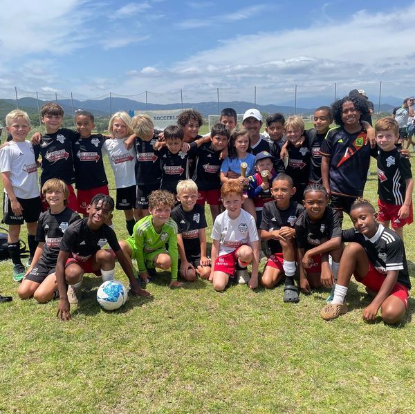 Youth soccer team posing on a grassy field with a ball under a partly cloudy sky