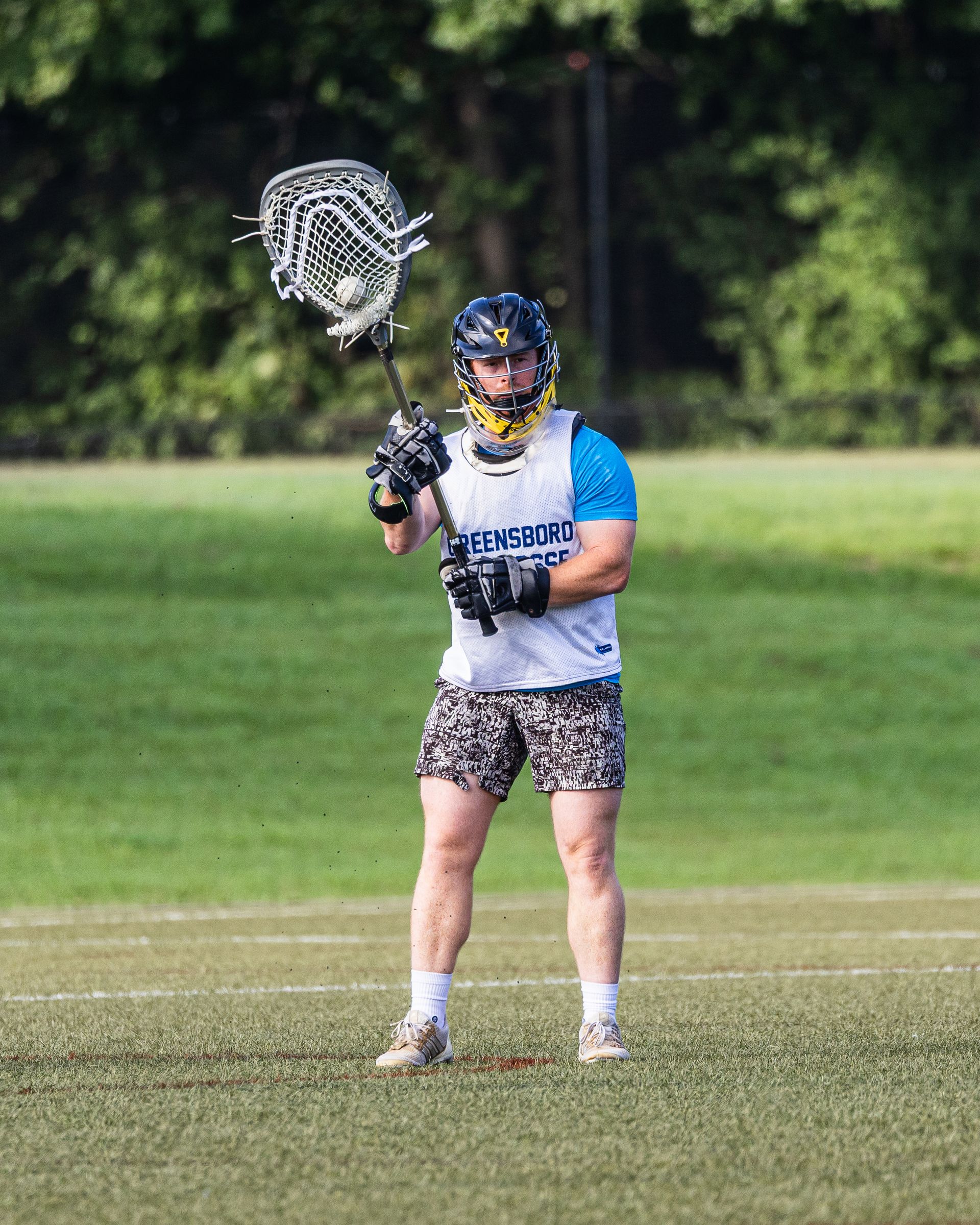 Lacrosse player in blue jersey and patterned shorts holding a stick on a grassy field
