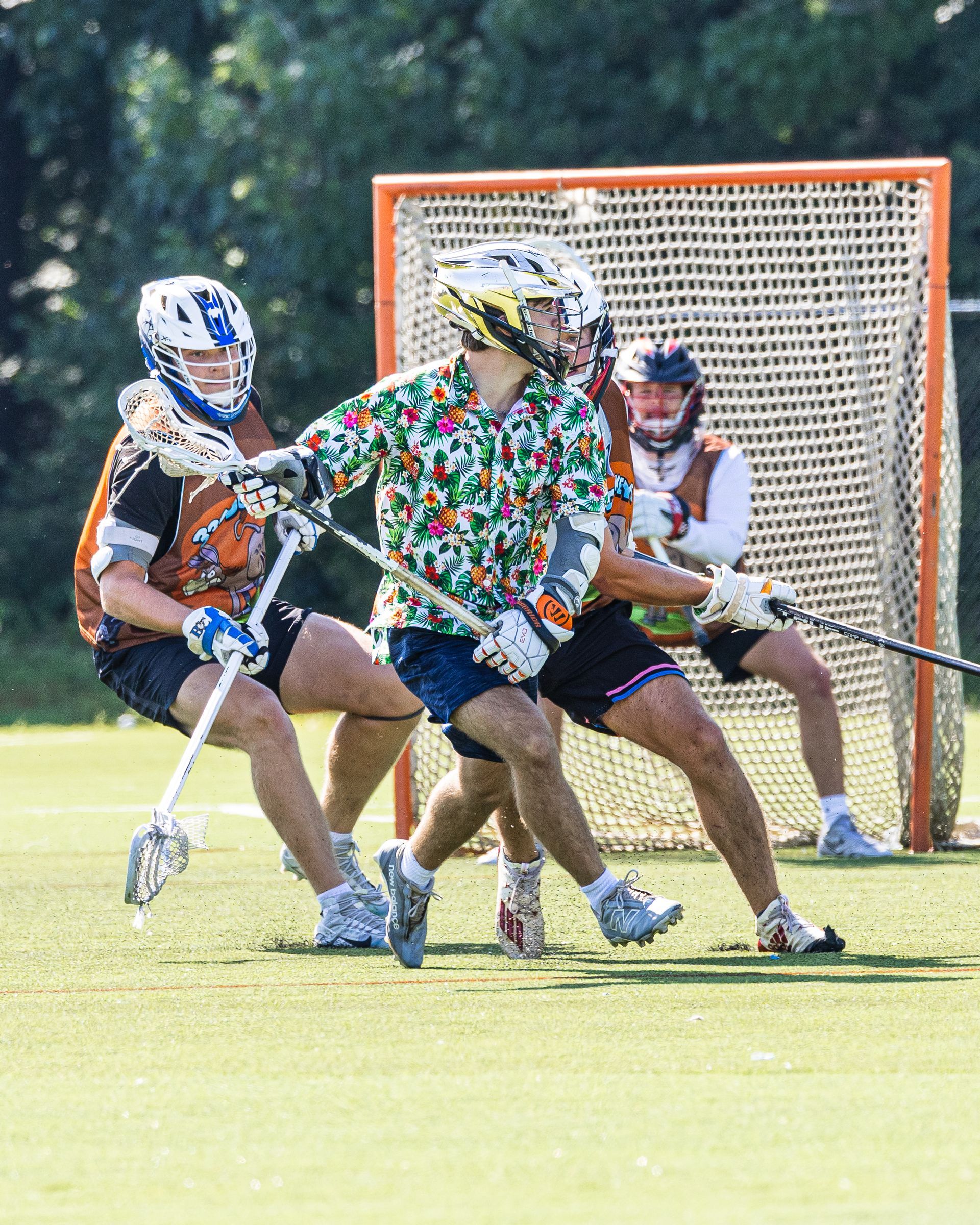 Three lacrosse players contest a ground ball in front of the goal on a sunny field.