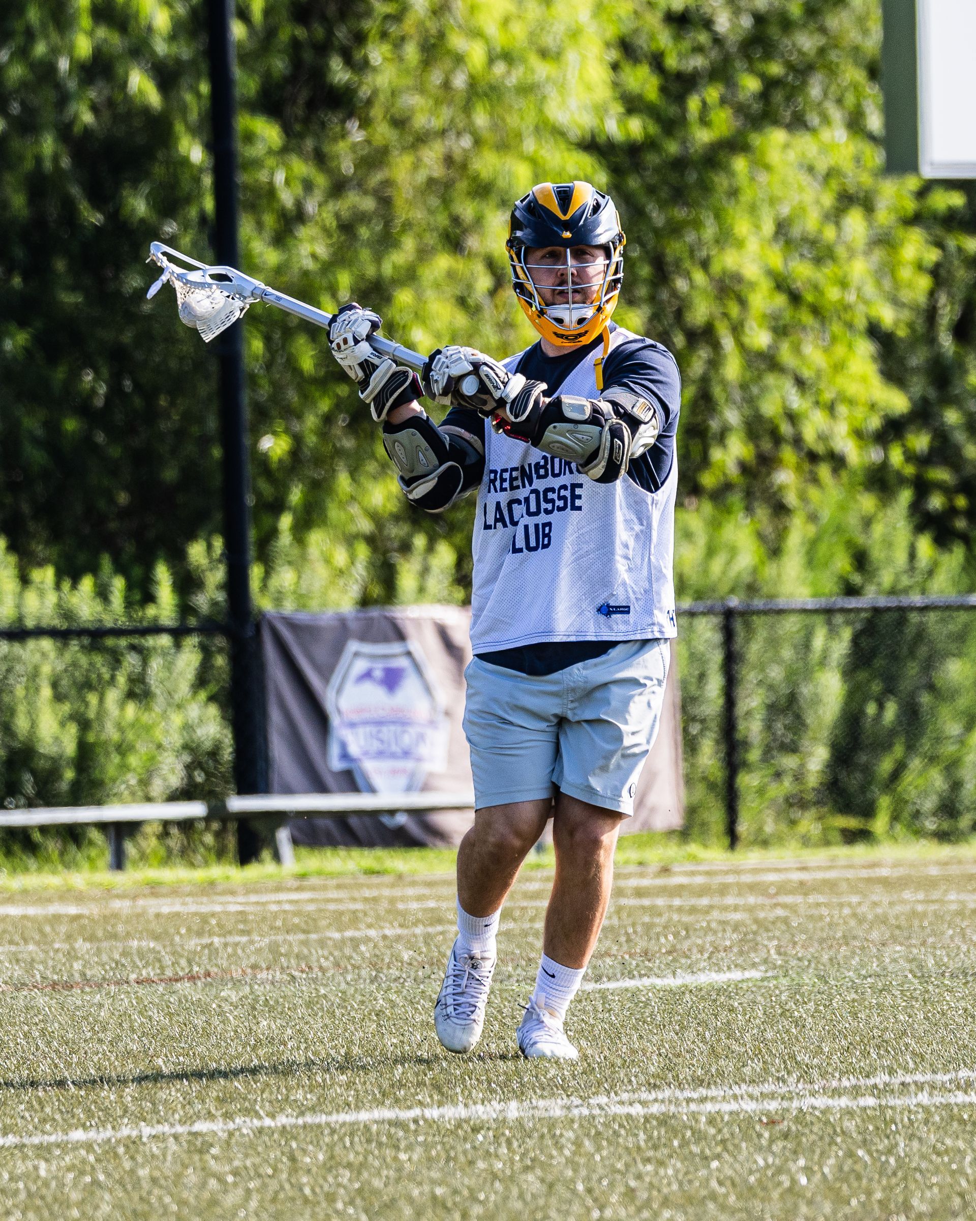 Lacrosse player in white uniform holds a stick on a grassy field, with trees and a fence in the background