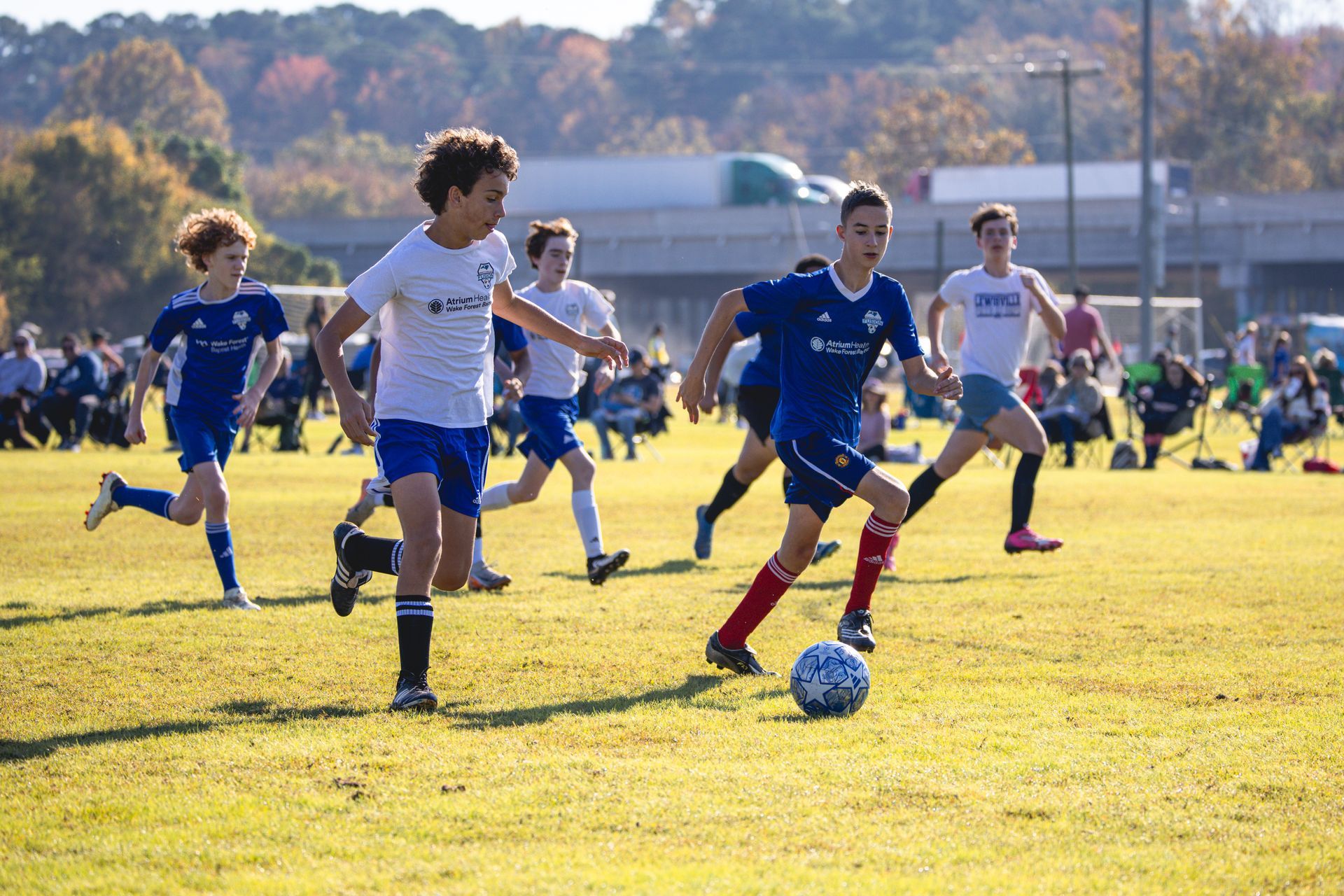 Youth soccer players in blue and white sprint across a sunny field, chasing the ball.