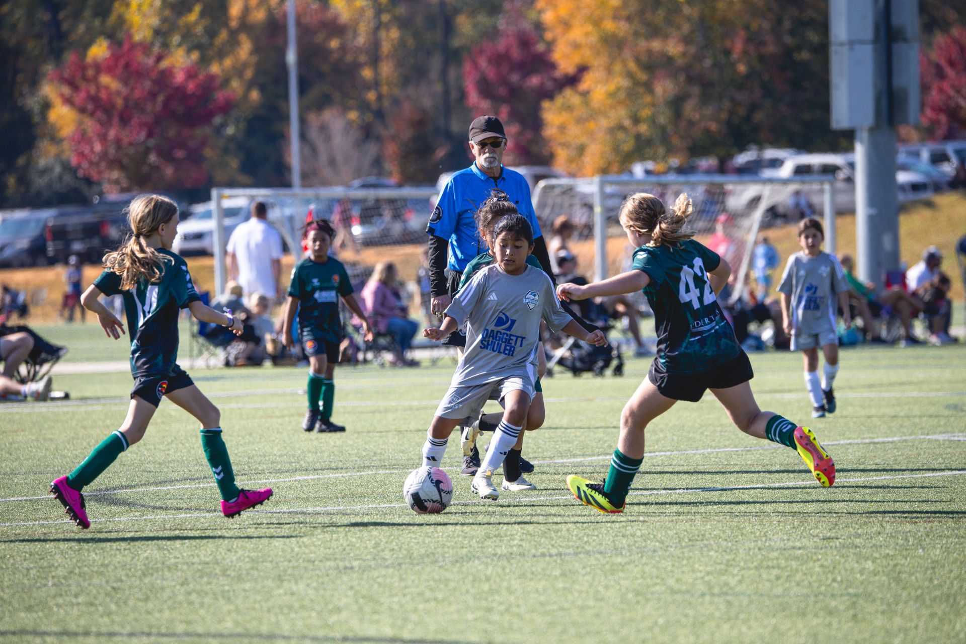 Youth soccer game with players chasing the ball on a grassy field, trees and spectators in the background