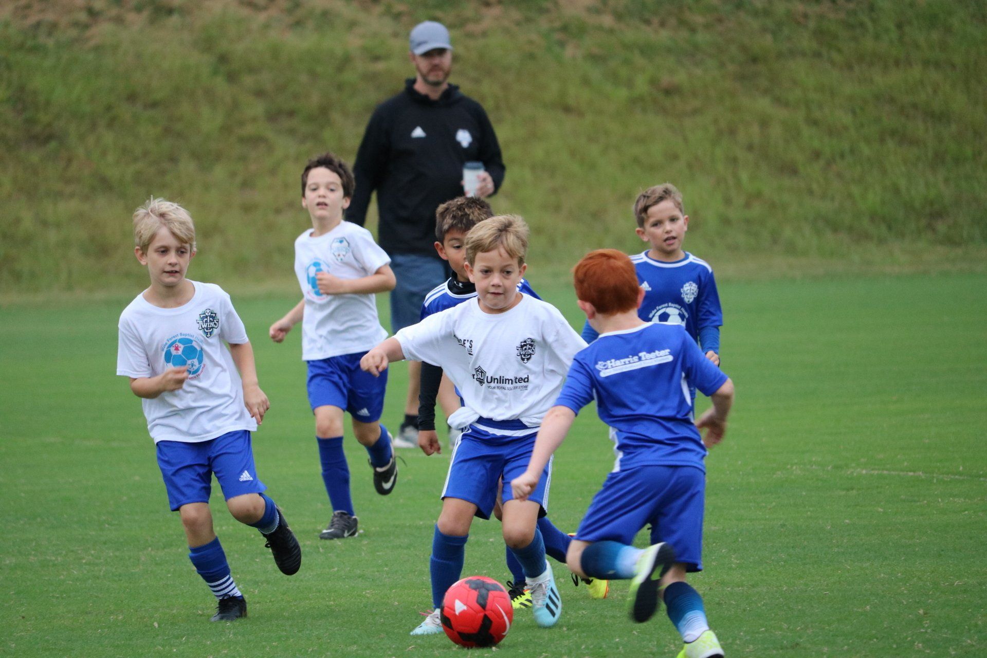 Children playing soccer on a grassy field, chasing a red ball with a coach in the background