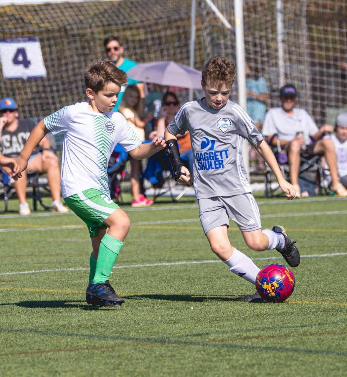 Two kids playing soccer, one in gray dribbling the ball while another in white and green defends on a field.