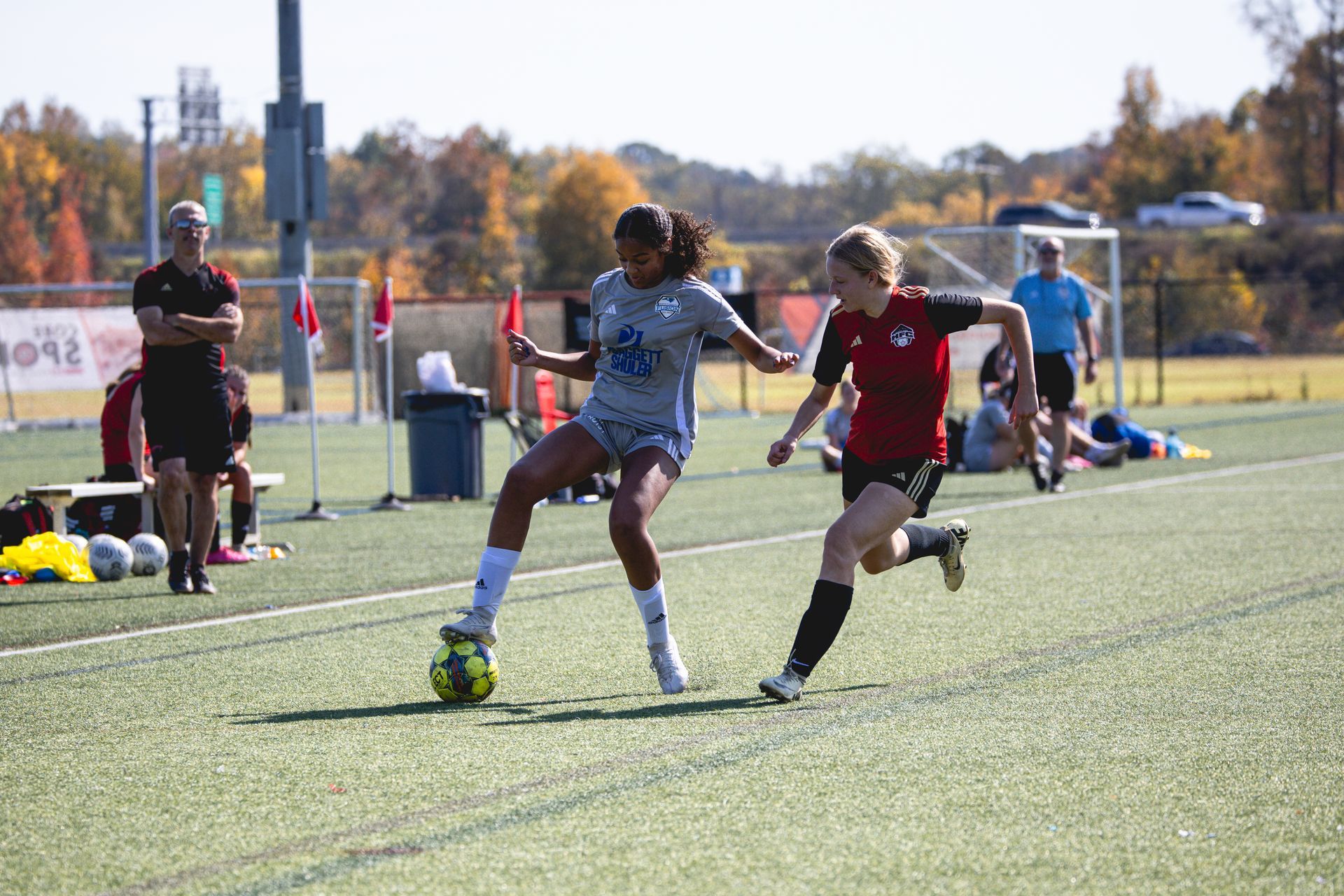 Two soccer players battle for the ball on a sunny field, with spectators and cones in the background.