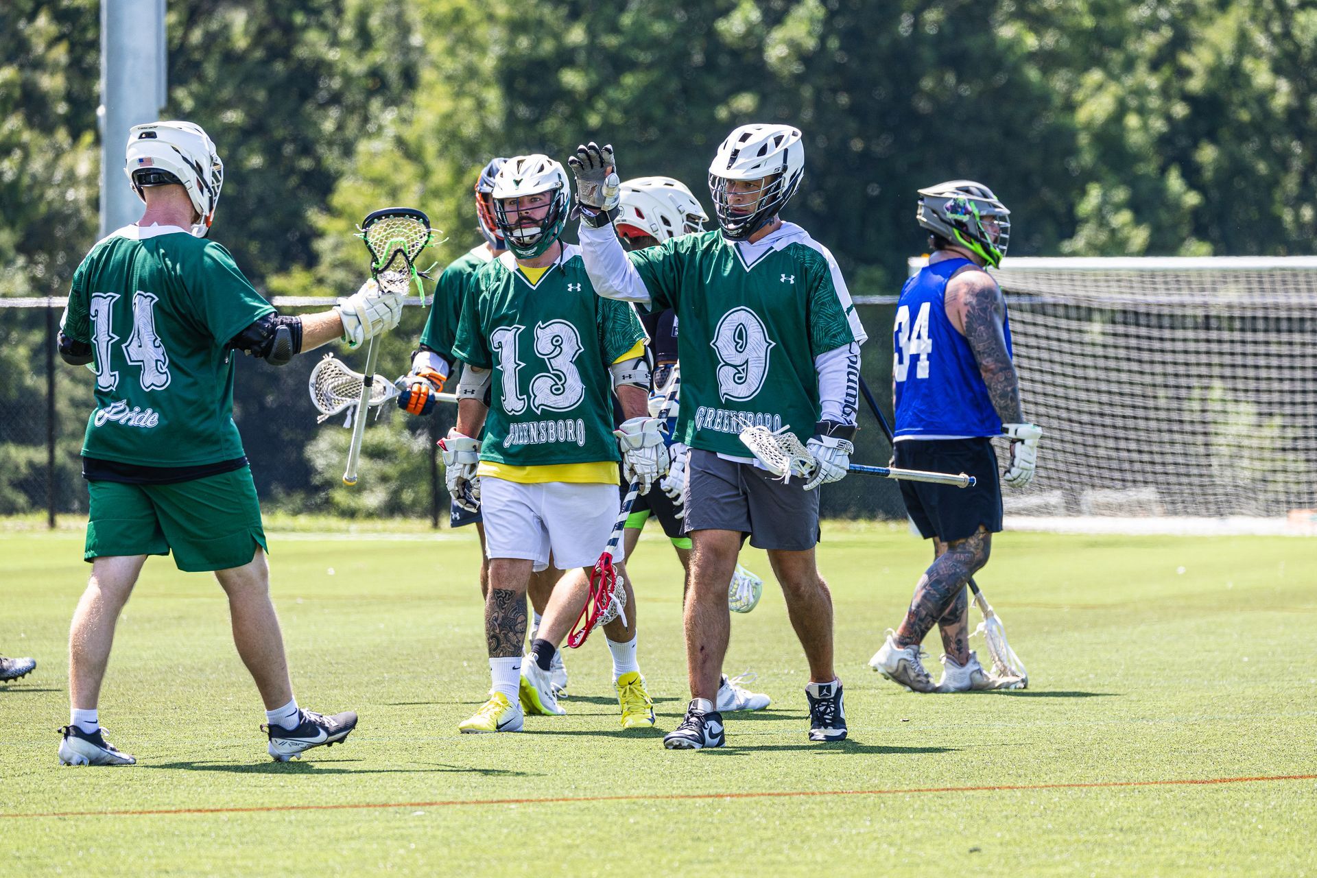 Lacrosse players in green and blue jerseys walking on a sunny field near a goal net