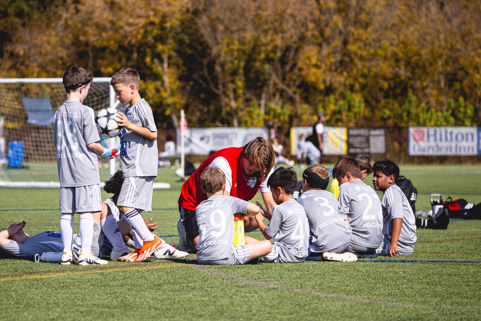 Soccer players in white uniforms huddle on a field while a coach in red talks to them.
