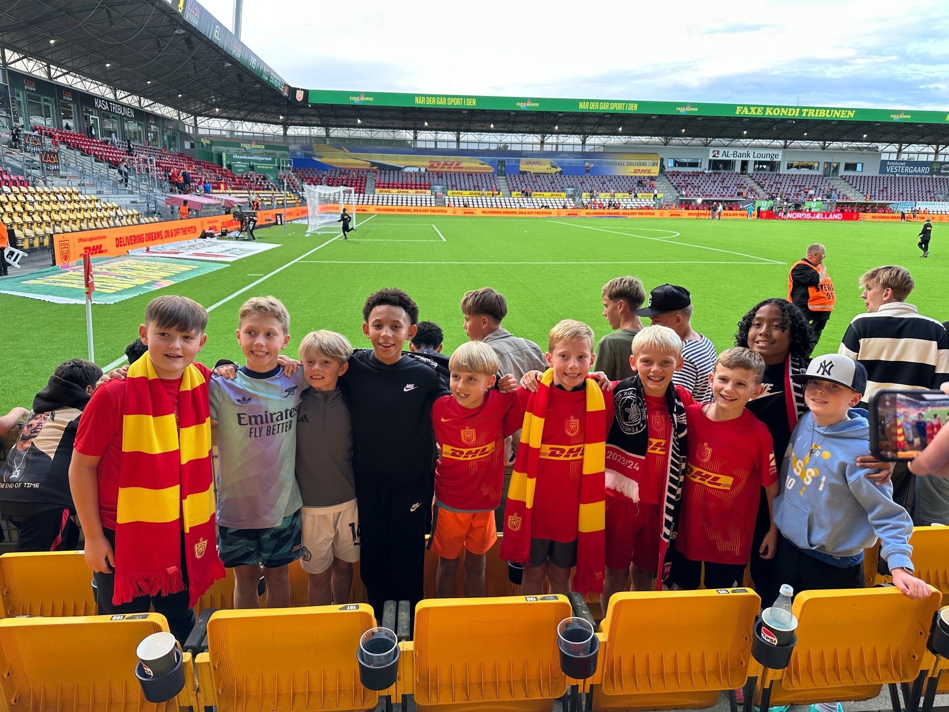 Children posing on a soccer field, several in red and yellow jerseys, with stadium seats in the background