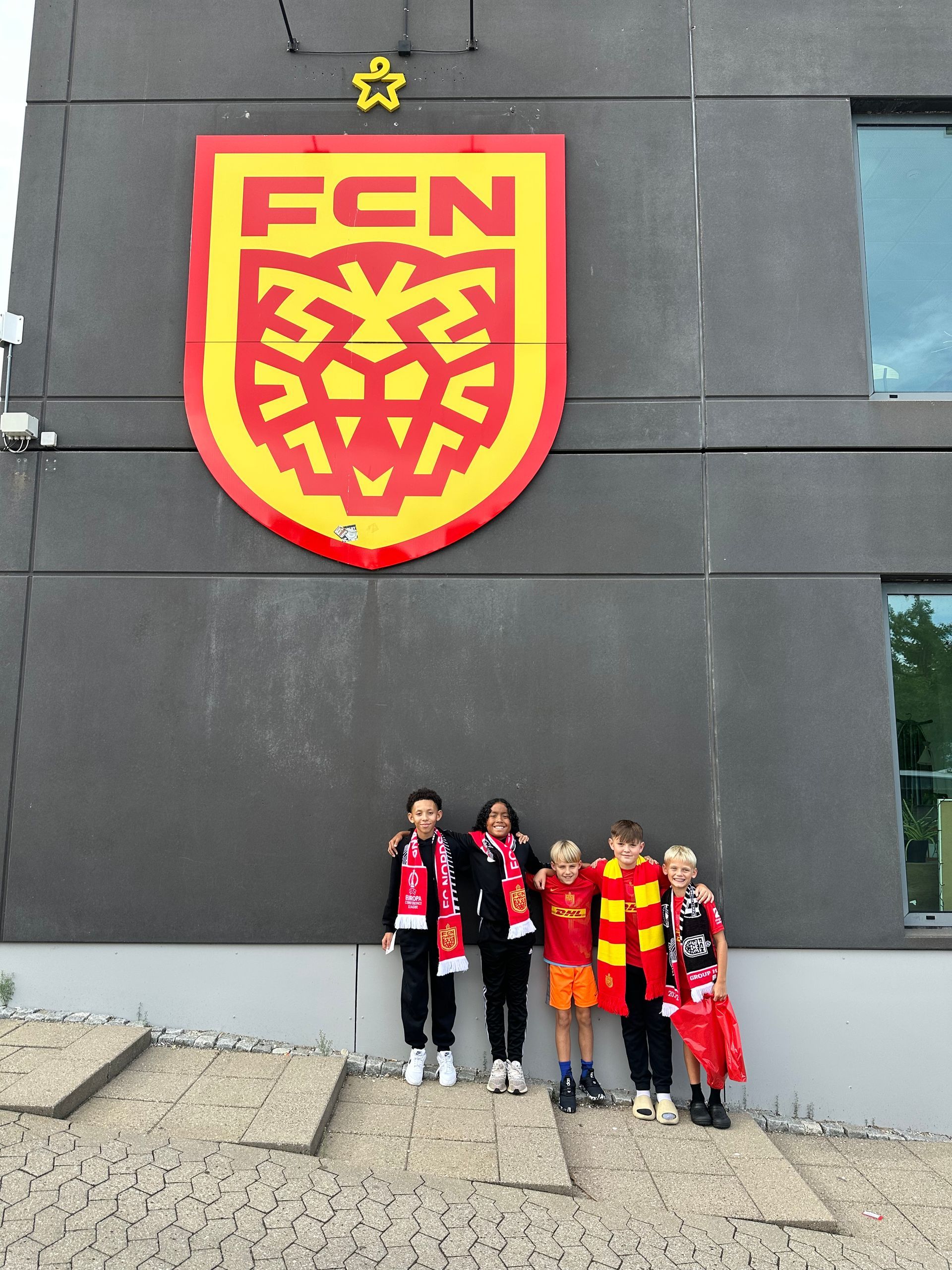 Group of soccer fans standing below a large FCN crest on a gray wall