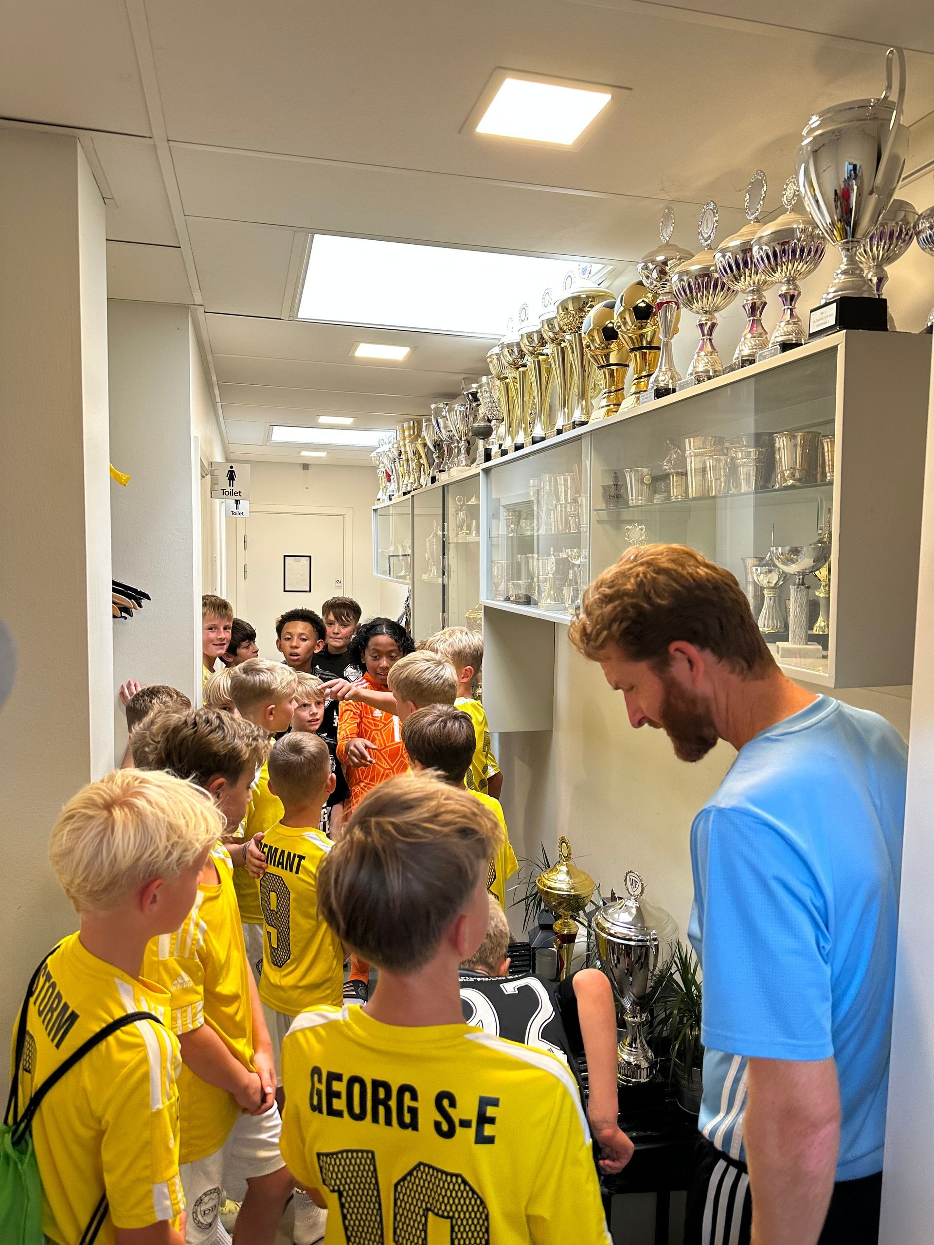 Soccer team in yellow jerseys crowded in a trophy-lined hallway, talking with a man in blue.