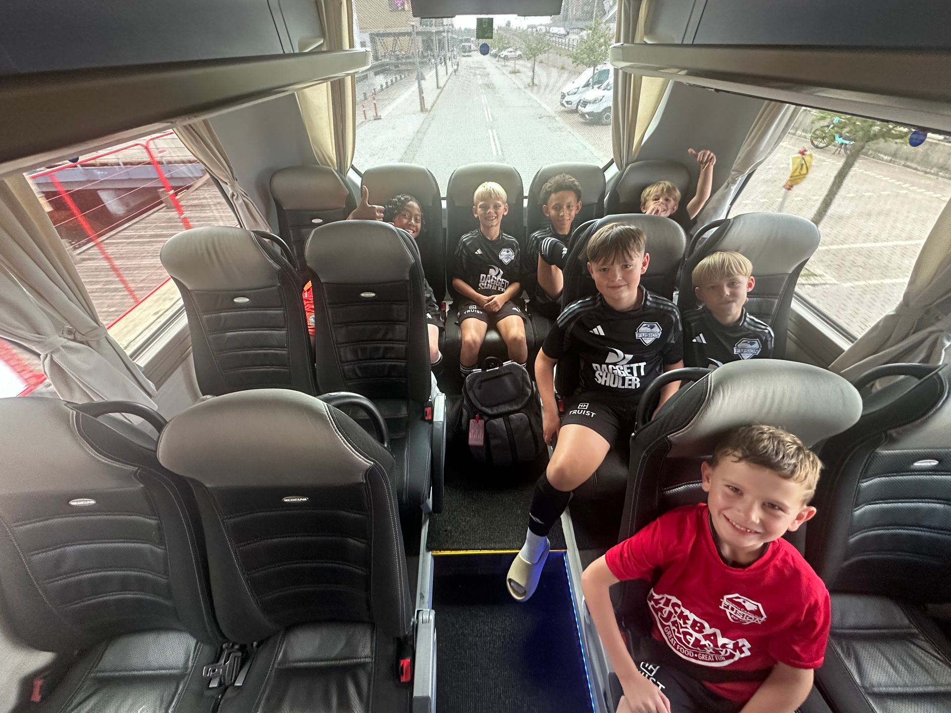 Several kids in black sports jerseys and one in red sit on a bus, smiling toward the camera.