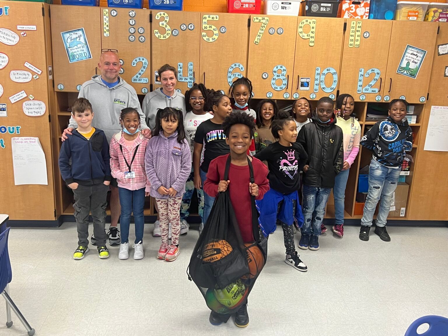 Group of children posing in a classroom, standing before a bulletin board decorated with colorful papers.