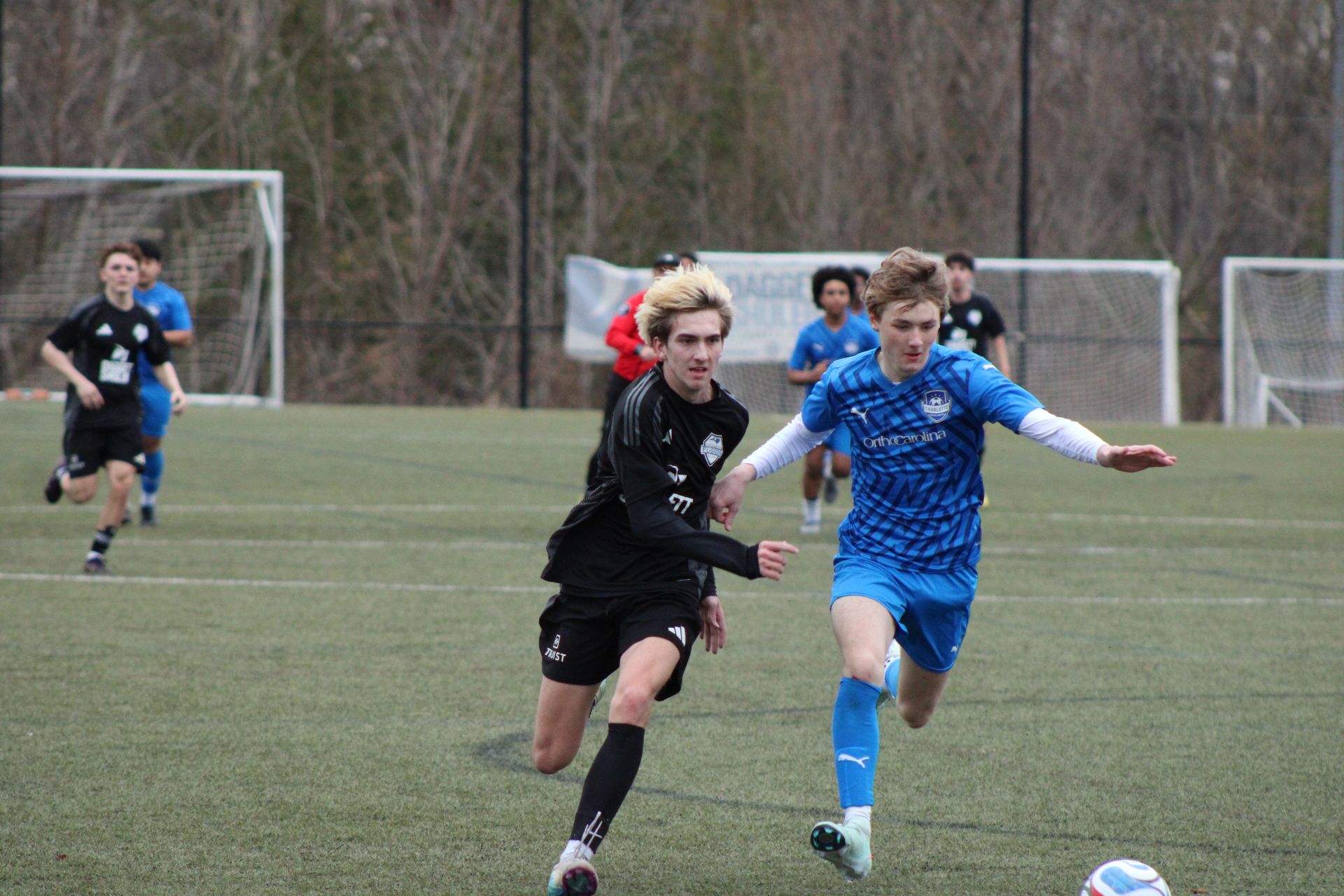 Two soccer players sprint after the ball on a field, with a goal and other players in the background.