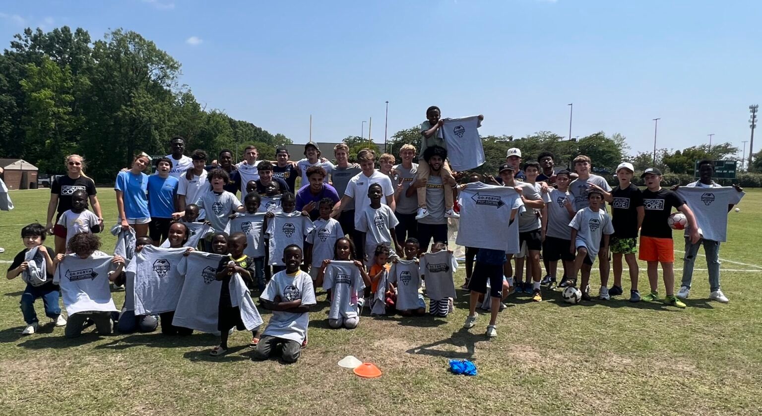 Large group posing outdoors on a sunny grassy field, many holding white signs and wearing matching shirts.
