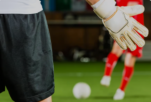 Goalkeeper in black shorts and white gloves on a soccer field, with a blurred player in red and a ball in the background