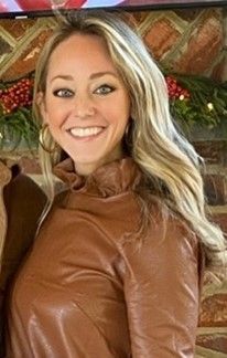 Smiling woman in a brown leather jacket standing indoors in front of a brick fireplace.