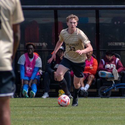 Soccer player in beige jersey dribbles the ball toward the camera on a field, with bench players behind him