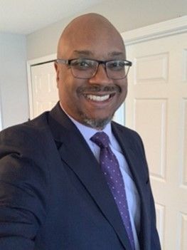 Smiling person in a navy suit and purple tie, standing indoors in front of white doors.