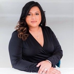 Portrait of a person seated at a desk, wearing a black wrap top against a light gray background.