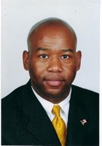 Formal portrait of a man in a black suit and yellow tie, facing forward with a neutral expression