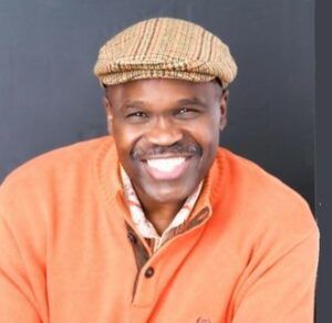 Smiling man in an orange sweater and plaid cap against a dark background