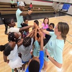 Children stack hands in a gym, with a coach guiding a team huddle on the court.