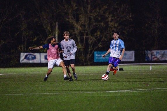 Three soccer players chase the ball on a lit field at night.
