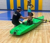 Man helping a person onto a bright green kayak in an indoor gymnasium