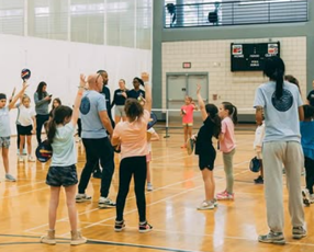 Children practicing volleyball drills in a gym with coaches overseeing from the court sides