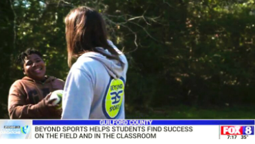 Two people talking outdoors near trees, with a news banner about sports helping students succeed.