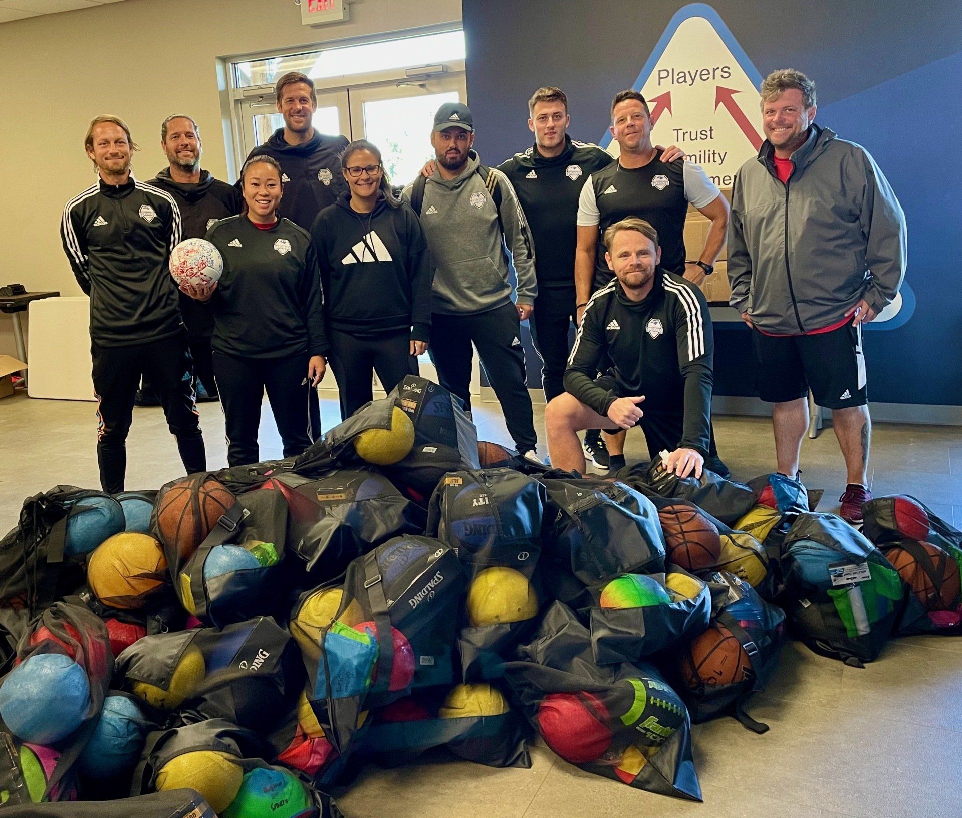 Group of people standing behind a large pile of colorful footballs indoors at a donation event