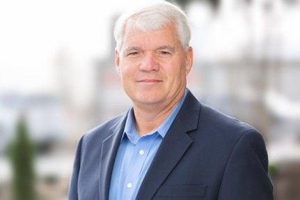 Smiling man in a navy blazer and blue shirt standing outdoors against a blurred background