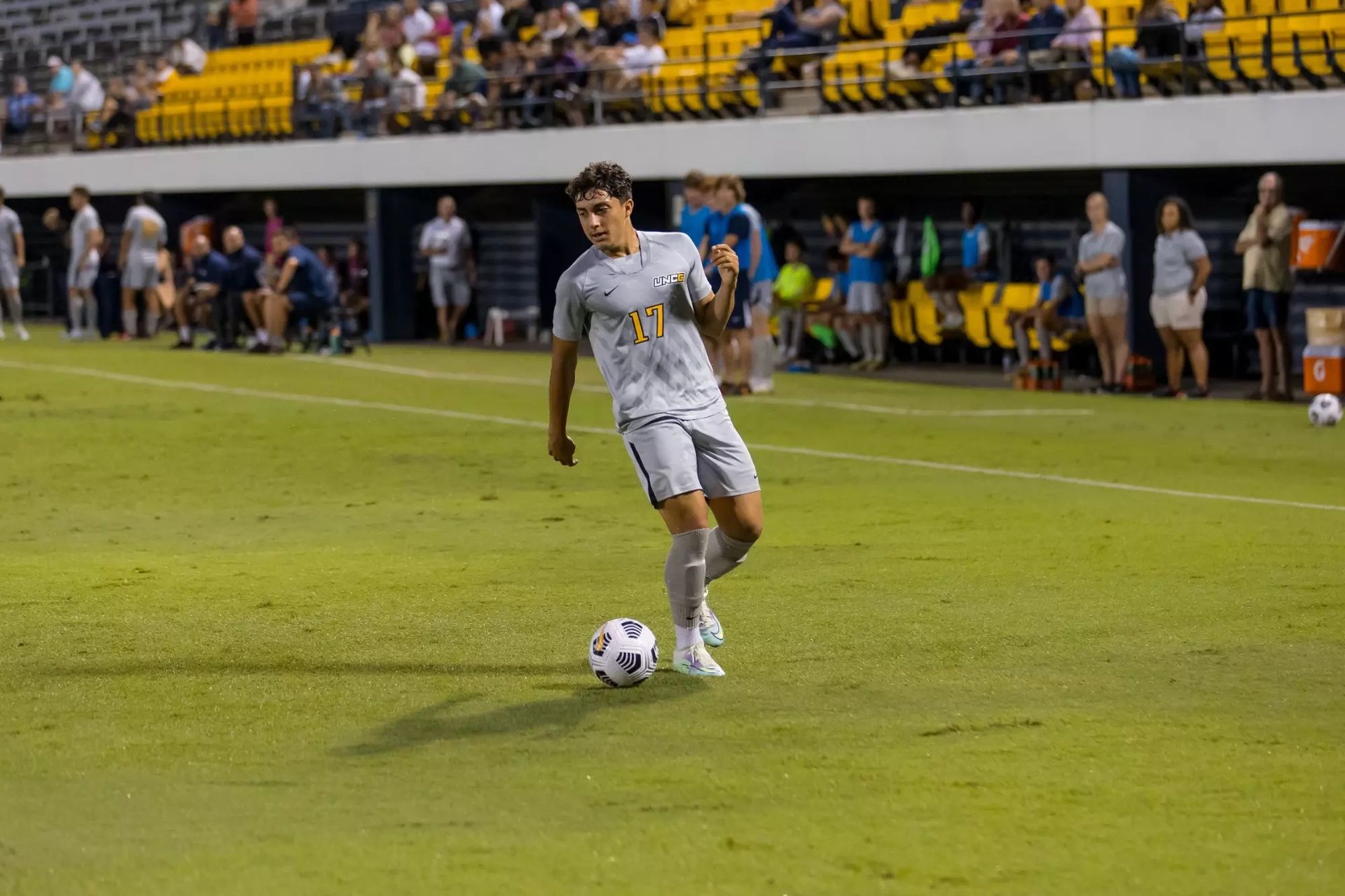 Soccer player in white dribbles the ball on a lit field with spectators and benches in the background