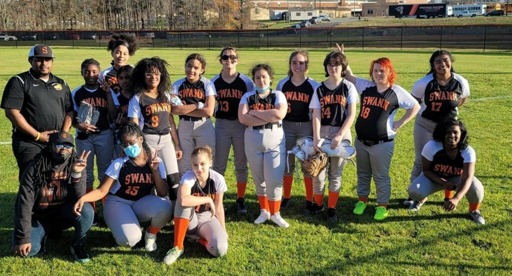 Youth softball team posing on a grassy field, wearing black jerseys with orange accents and white pants.