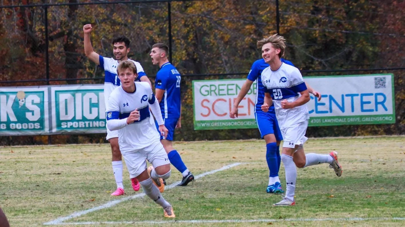 Soccer players in white and blue uniforms sprint on a muddy field during a match