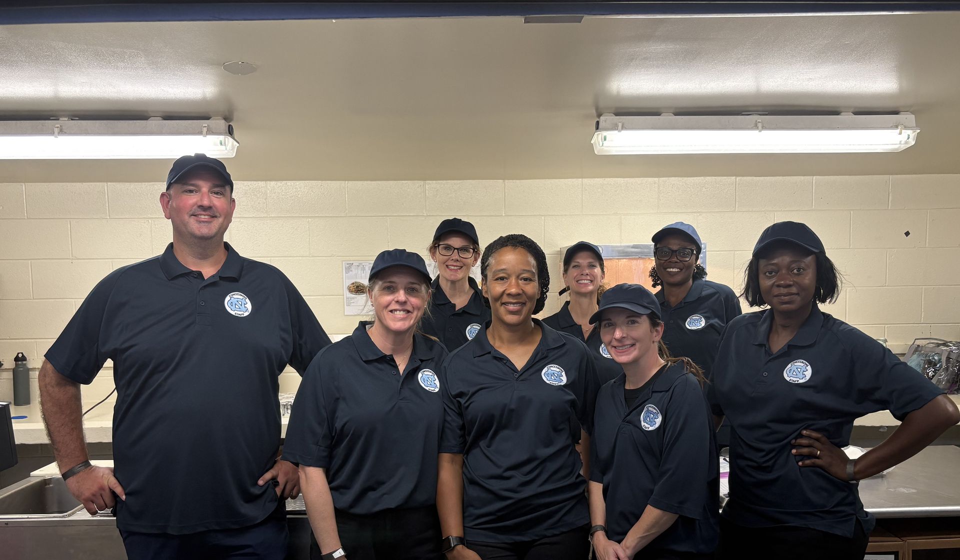 Seven people in black shirts posing indoors, smiling in a group photo under fluorescent lights