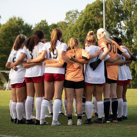 Soccer team huddling on the field, seen from behind, in white uniforms with red shorts during sunset