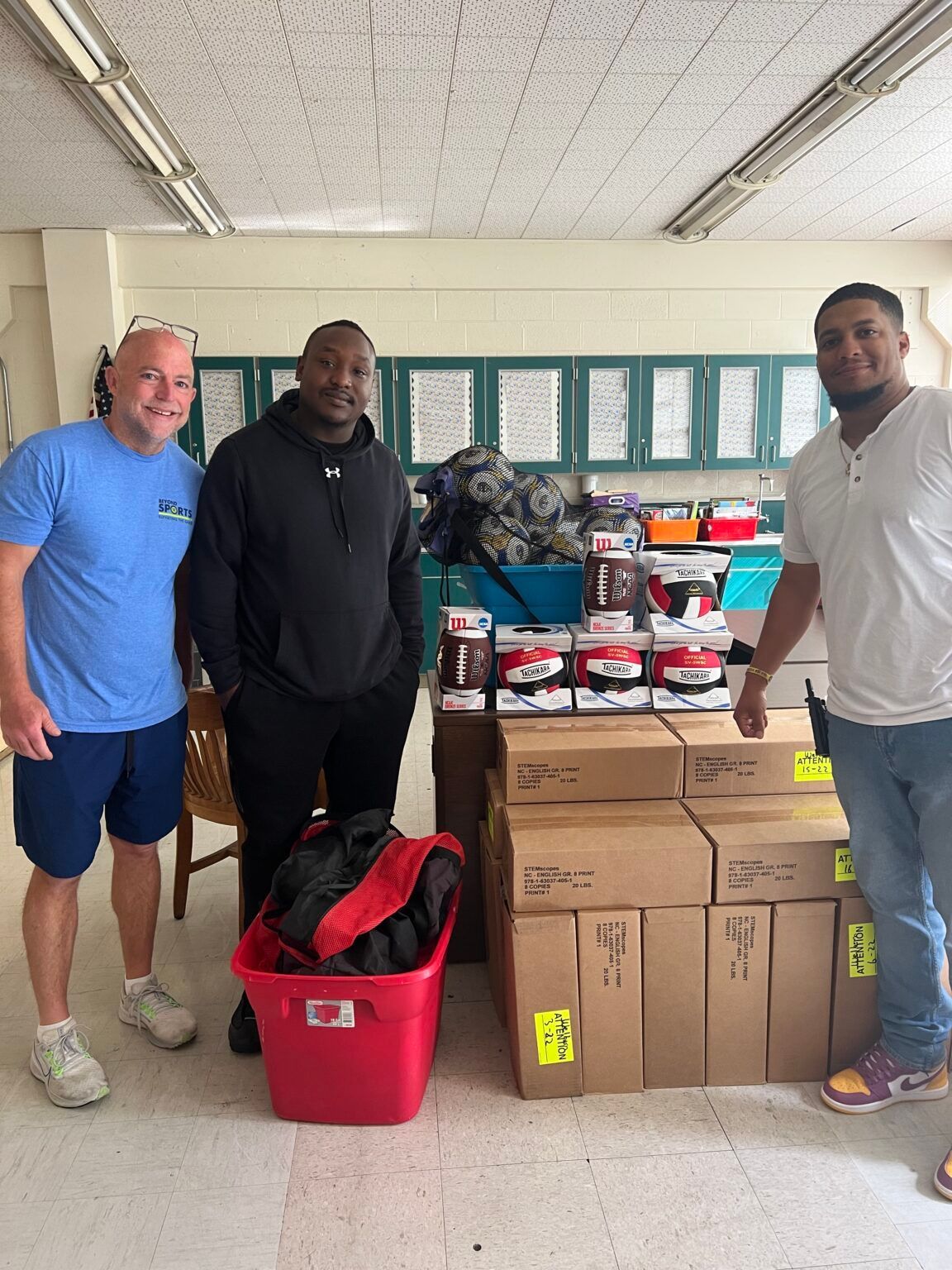 Three men stand beside a donation pile of shoe boxes and a red bin in a room.