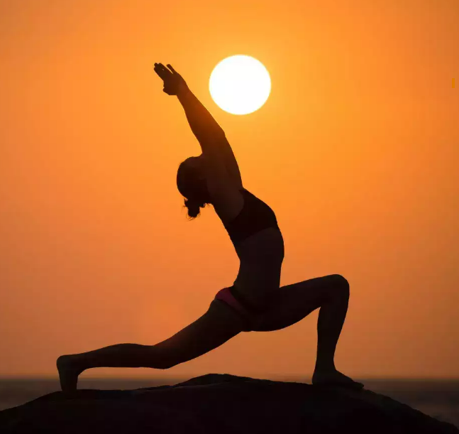 Silhouette of a person in a yoga lunge pose against an orange sunset sky with the sun behind them