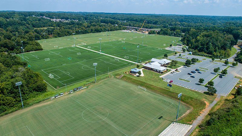 Aerial view of a green sports field with white markings, nearby parking lot, and trees surrounding the complex.