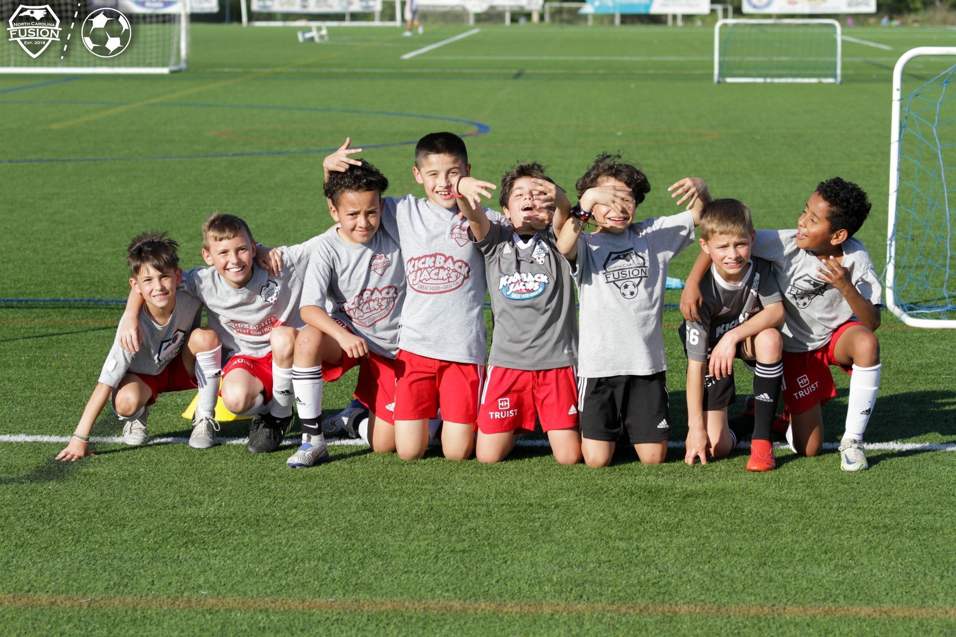 Youth soccer team posing on a grassy field, kneeling in gray jerseys and red shorts with goals in the background