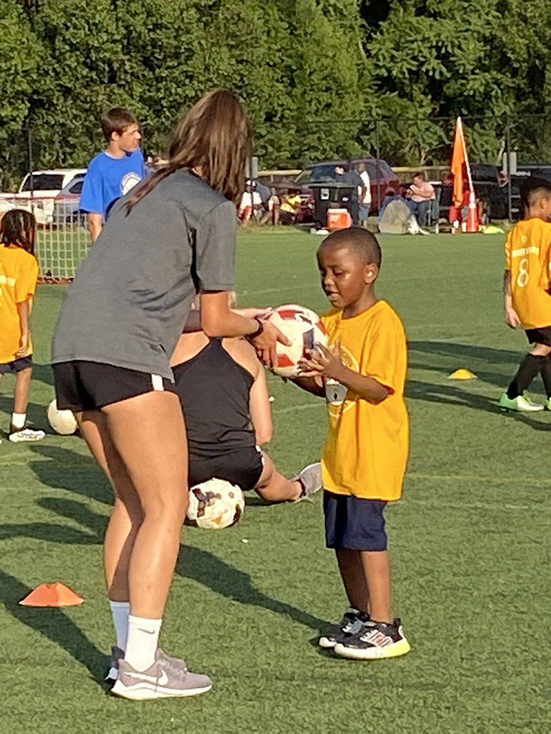 Soccer coach kneels with child holding a ball during youth practice on a grassy field.