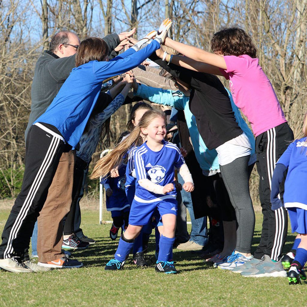 Children in blue sports uniforms run through a tunnel of raised arms on a grassy field.