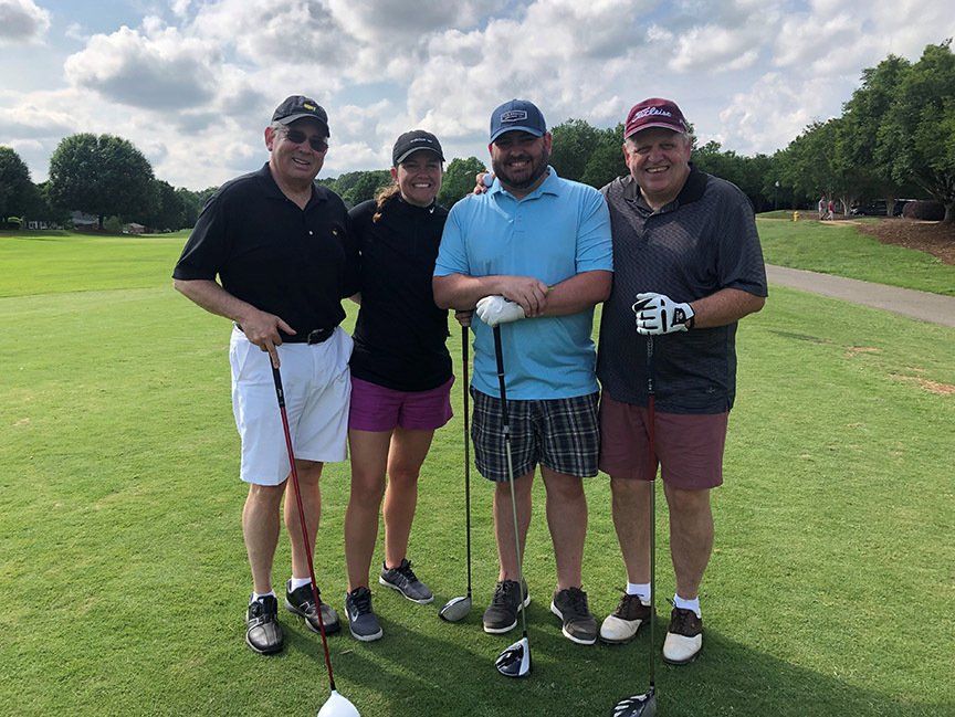 Four golfers posing on a green fairway, holding clubs under a cloudy sky.