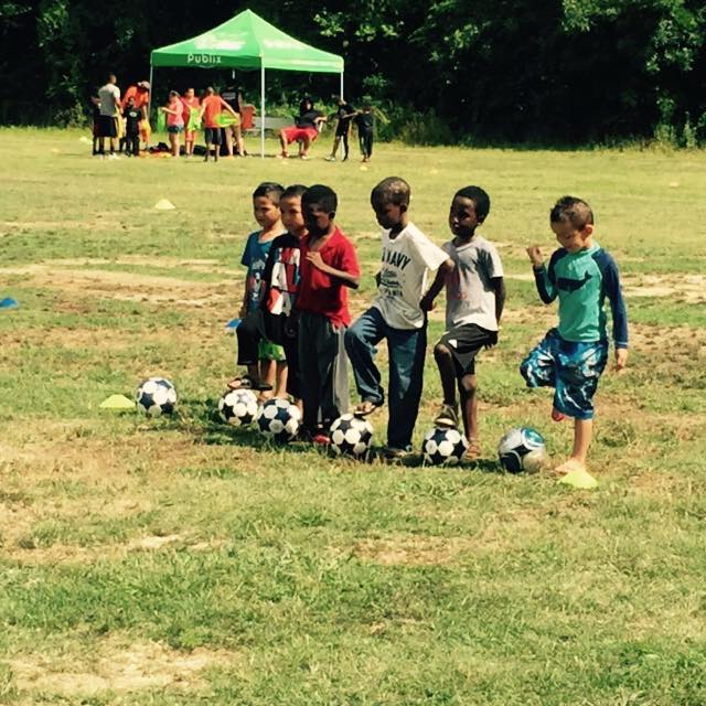Children standing by soccer balls on a grassy field, with a green tent and people in the background.