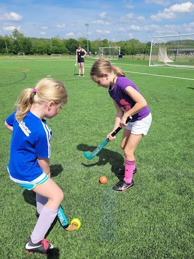 Two children practicing field hockey on a turf field with cones and a goal in the background