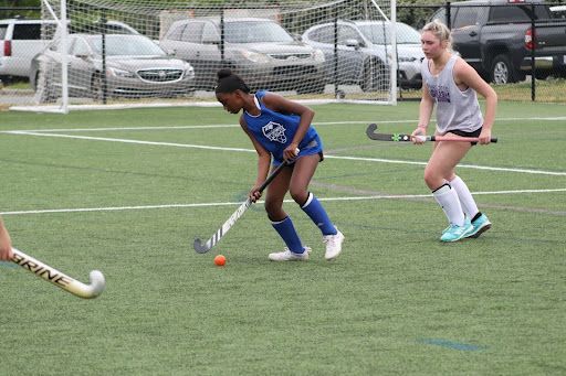Field hockey players chase an orange ball on an outdoor turf field.